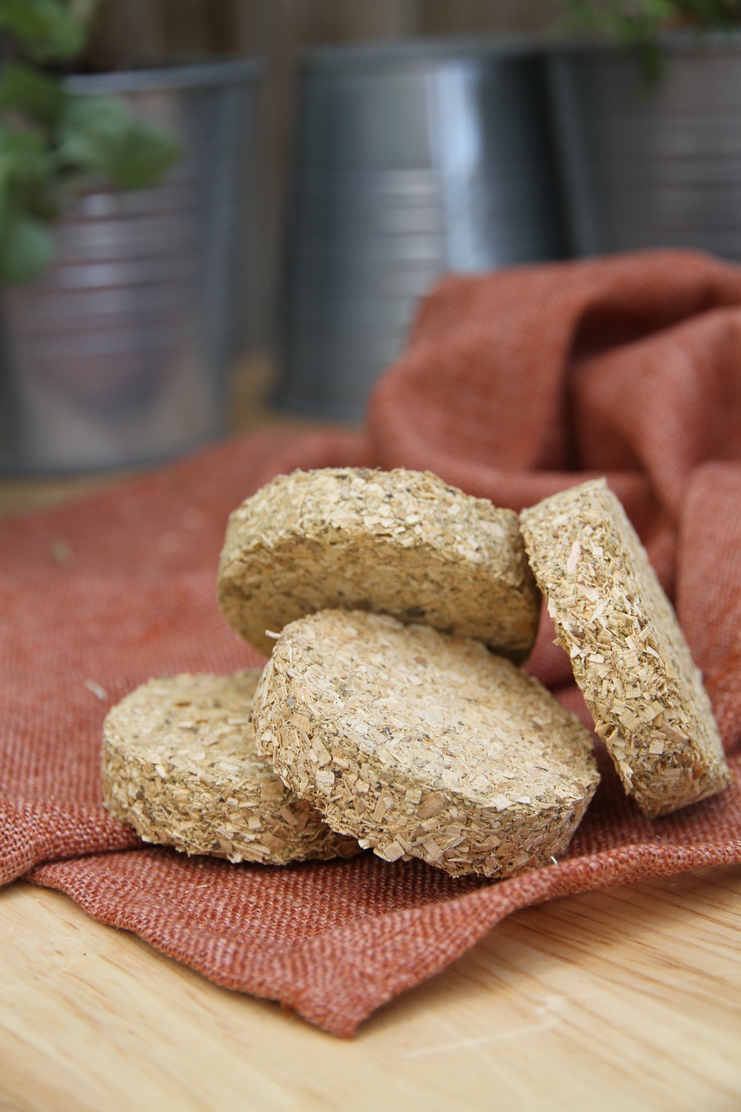 Four light-colored Bradley Smoker Bisquettes made of compressed wood, arranged on a rust-colored linen napkin, with blurred metal pots of assorted herbs in the background.